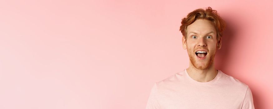 Headshot of amazed redhead male model in t-shirt open mouth, looking fascinated and happy at camera, standing over pink background.