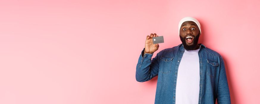 Shopping concept. Amazed Black guy showing credit card, staring at camera impressed, recommending bank, standing over pink background.