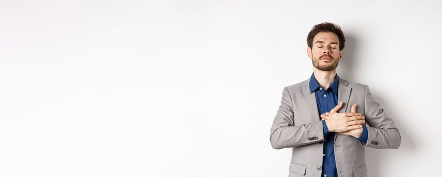 Young man in suit close eyes, holding hands on heart and remember happy memories, feeling nostalgic, standing against white background.