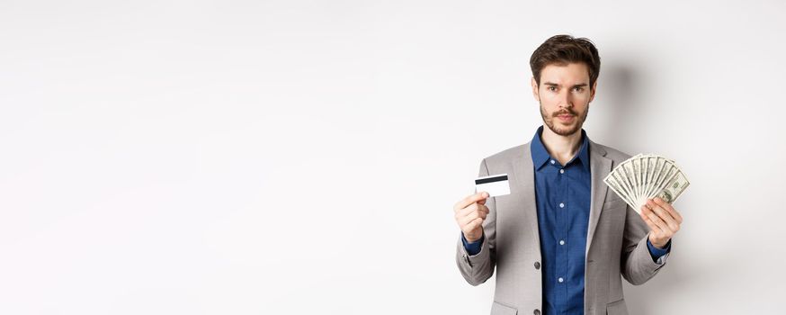 Successful businessman making money, standing in suit with dollar bills and plastic credit card, white background.