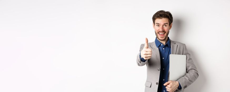 Good job. Happy smiling business man in suit holding laptop, showing thumb up to praise you, nice work gesture, standing on white background.