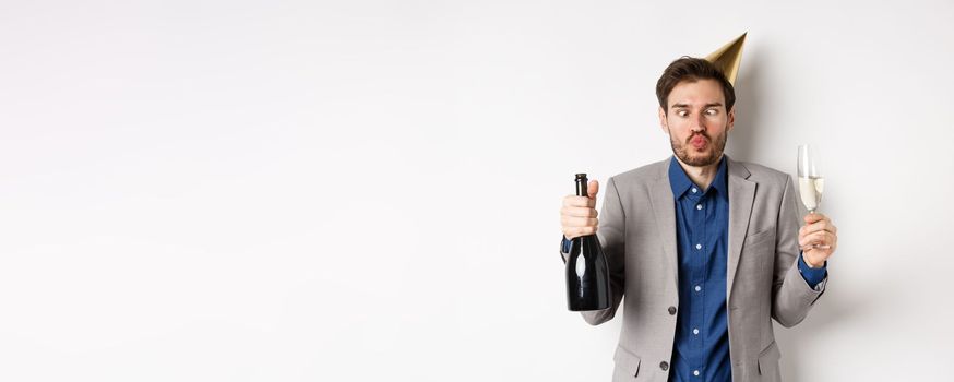 Celebration and holidays concept. Funny drunk guy in suit and birthday hat, squinting eyes and having fun at party, drinking champagne, white background.