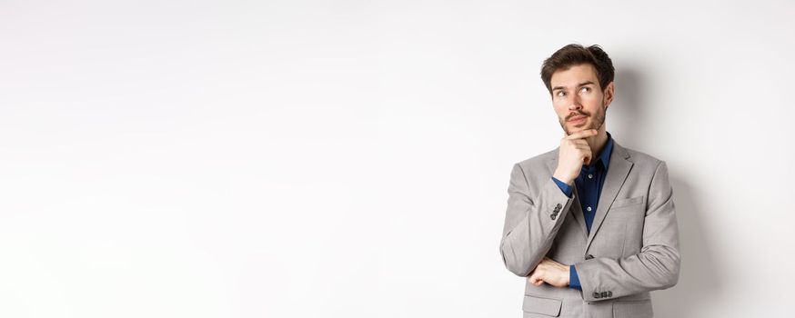 Pensive handsome man in suit looking up and thinking, making choice, pondering ideas, standing against white background.