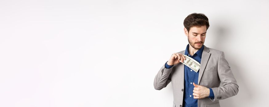 Successful businessman put dollars inside suit pocket, hiding money, standing on white background.