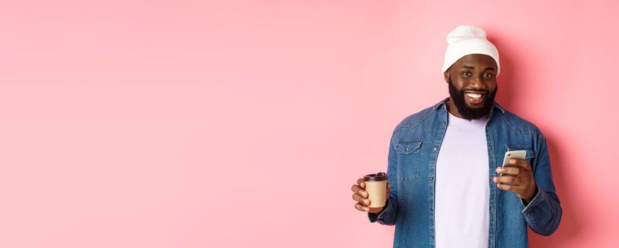 Image of stylish Black man hipster drinking takeaway coffee, reading message on phone and smiling, standing over pink background.