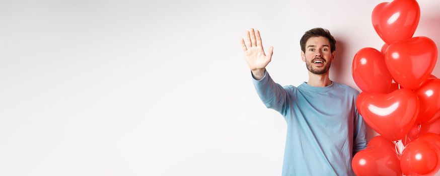 Handsome young caucasian man standing with romantic heart balloon and waving hand at lover, waiting for his date on Valentines day with cute surprise, standing over white background.