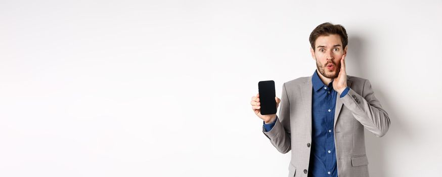 E-commerce and online shopping concept. Excited man gasping amaze and showing empty smartphone screen, wearing business suit, white background.