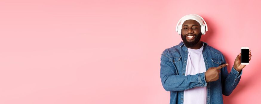 Happy black man listening music in headphones and smiling, pointing mobile phone screen app or playlist, standing over pink background.