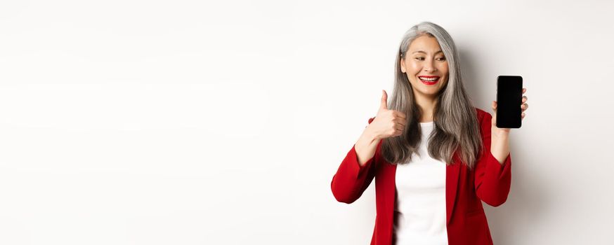 Satisfied asian elderly businesswoman showing blank smartphone screen and thumb-up, praising online promotion or company app, standing over white background.