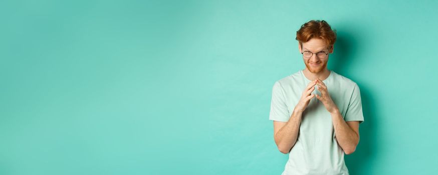 Devious redhead man in glasses and t-shirt pitching an idea, steeple fingers and look from under forehead with sly and smug smile, standing over mint background.