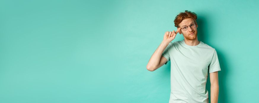 Skeptical redhead man scolding someone stupid or crazy, pointing finger at head and staring at camera, standing over mint background.