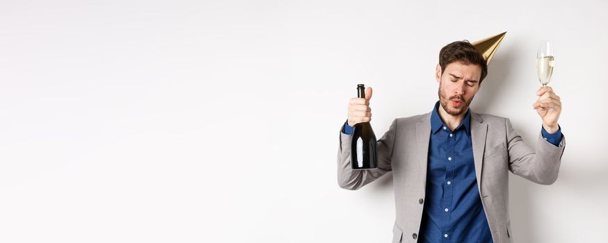 Celebration and holidays concept. Drunk guy dancing at party in birthday hat and suit, raising glass of champagne, having fun at event, white background.