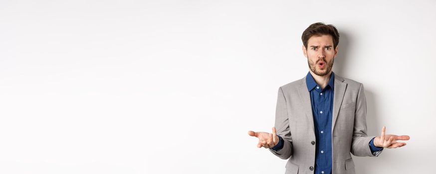 Confused and shocked businessman raising hands up and asking what happened, frowning disappointed, waiting for explanation, standing in suit on white background.