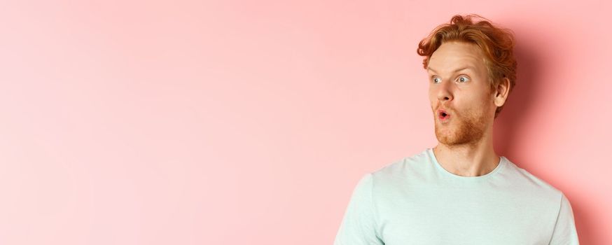 Headshot portrait of surprised redhead man with beard, looking left and saying wow, raising eyebrows amazed, standing over pink background.