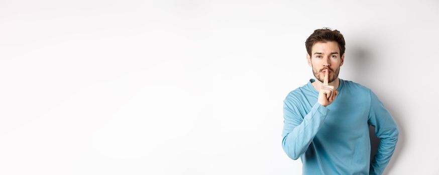 Attractive bearded man asking to keep quiet, showing taboo hush gesture and looking at camera calm, standing over white background.