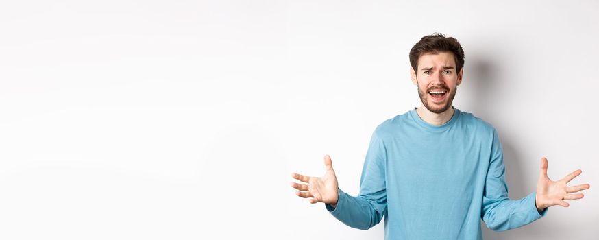 Image of frustrated and worried young man shaking hands, trying to explain something, standing anxious against white background.