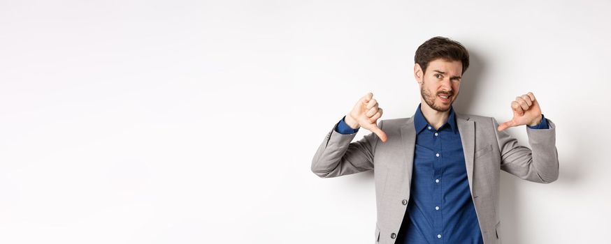 It sucks. Man feeling awkward showing thumbs down and judging something bad, frowning disappointed, standing in suit on white background.