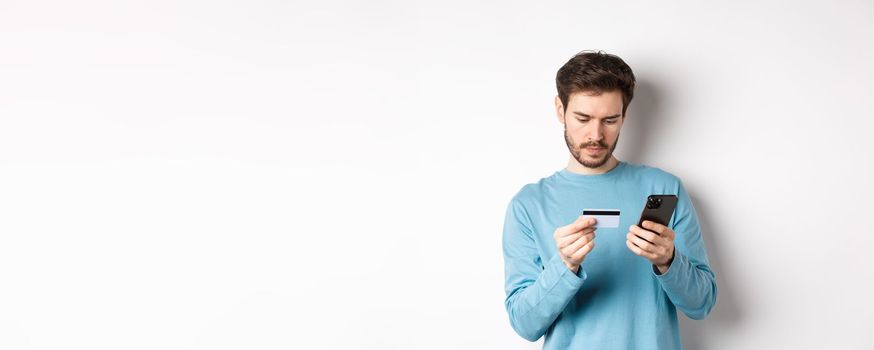 E-commerce and shopping concept. Young man making online payment, holding plastic credit card and smartphone, standing on white background.