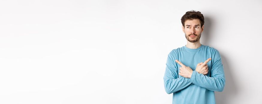 Indecisive man making choice, pointing fingers sideways at two variants and deciding, looking pensive at left side, standing on white background.