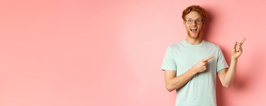 Amazed redhead man in glasses and t-shirt showing special promotion, pointing fingers at upper right corner and staring excited at camera, standing over pink background.