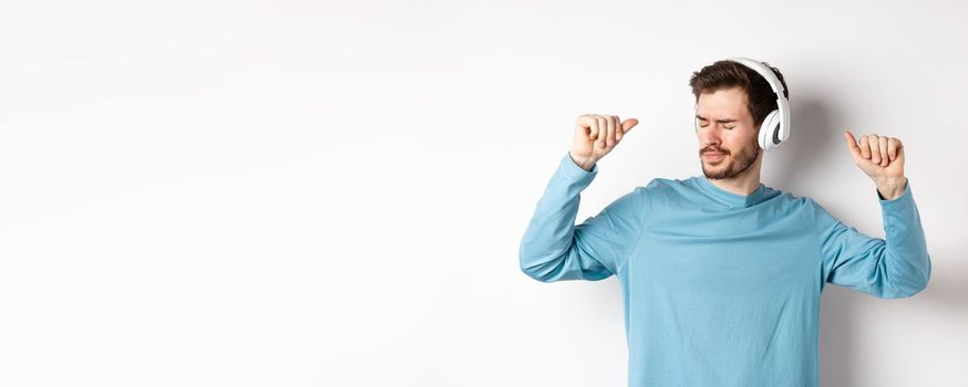 Happy young man having fun in headphones, dancing while listening music in wireless earphones, white background.