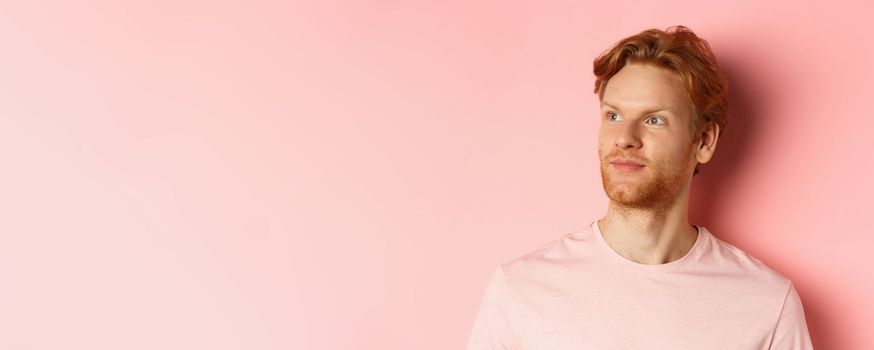 Handsome european male model with red hair and beard, turn head and looking pleased at copy space on left side, standing over pink background.