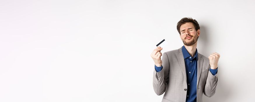 Excited successful businessman dancing with plastic credit card, raising fist pumps and smiling delighted, standing on white background in suit.