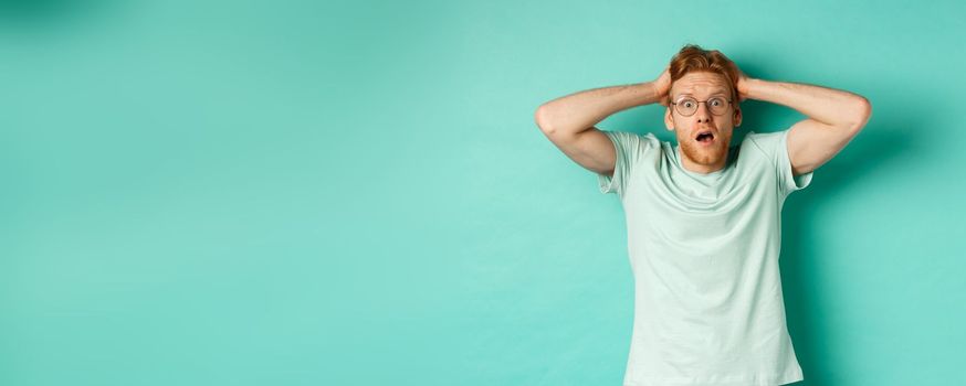 Young redhead man with beard, wearing glasses and t-shirt, holding hands on head and staring in panic, standing alarmed and anxious against turquoise background.