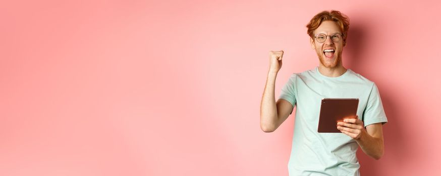 Image of happy redhead man triumphing, winning online with digital tablet and rejoicing, standing over pink background.