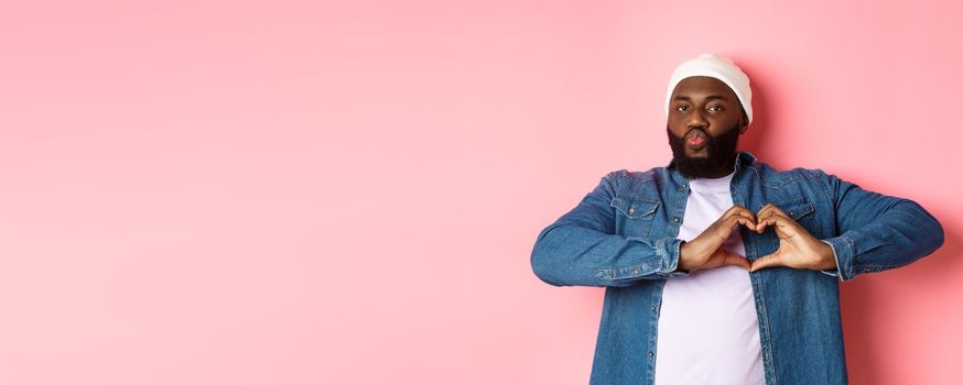 Happy african-american man showing heart sign, I love you gesture, pucker lips for kiss while standing over pink background.