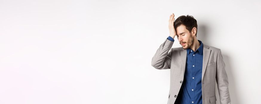 Annoyed businessman in suit facepalm with eyes closed, bothered by someone stupid, pissed-off at work, standing on white background.