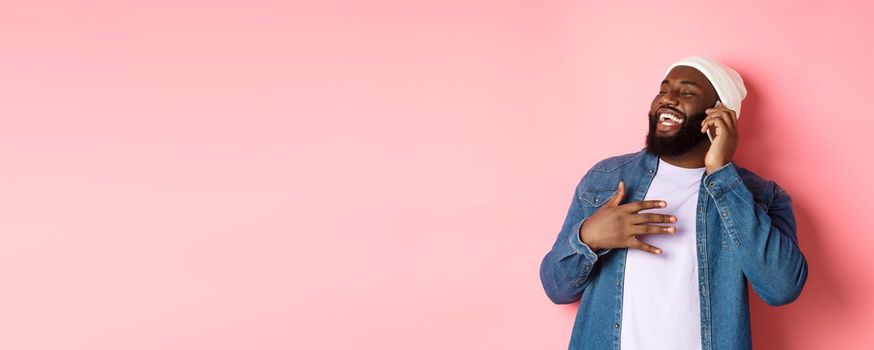 Happy african-american man talking on mobile phone, laughing and smiling, standing in beanie and denim shirt over pink background.