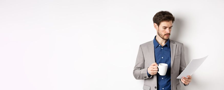 Boss reading documents and drinking morning coffee from mug, looking at paper serious, standing in suit on white background.