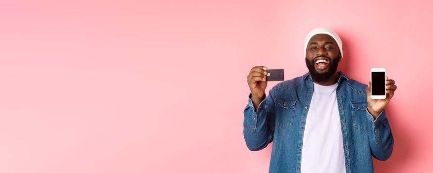 Online shopping. Happy Black man laughing and smiling, showing credit card and smartphone screen, recommending phone app, pink background.