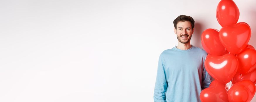 Smiling young man standing with heart balloons and looking happy, celebrating valentines day, bring romantic present to lover, standing over white background.