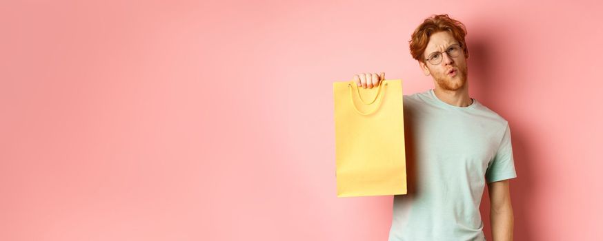 Handsome young man in glasses looking sassy at camera, showing yellow shopping bag, telling about discounts in shop, standing over pink background.