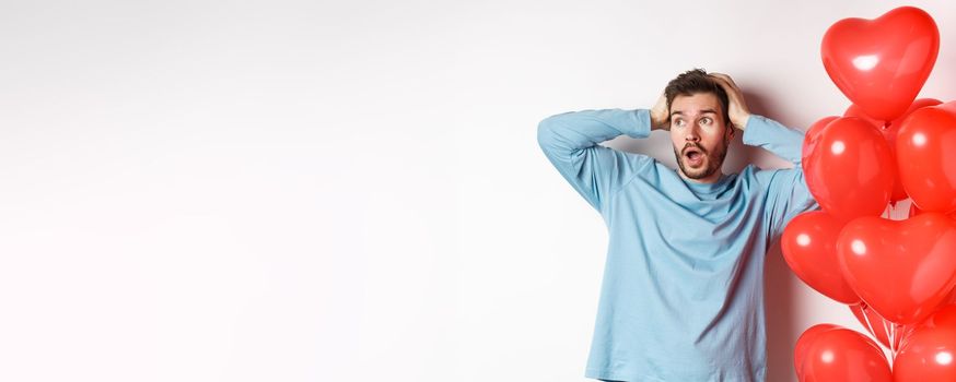 Boyfriend grab head in hands and panic over Valentines day gifts, looking sideways with alarmed face, standing near hearts balloons and thinking of present for lover, white background.