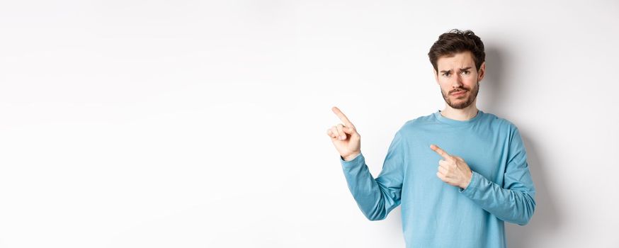 Skeptical young man with beard grimacing unsatisfied, frowning and pointing at upper left corner logo doubtful, standing upset over white background.