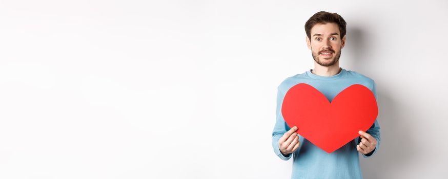 Relationship and love concept. Handsome caucasian man in sweater holding big red valentines day heart cutout and smiling, confessing on date, standing over white background.