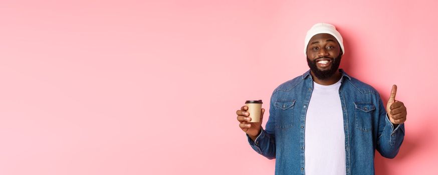 Handsome african-american hipster man showing thumb up, drinking coffee and recommending cafe, standing over pink background.