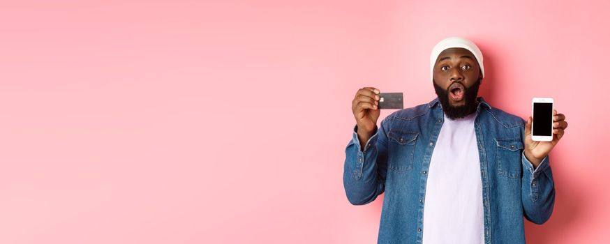 Online shopping. Excited Black man showing credit card and mobile phone screen, standing over pink background amazed.