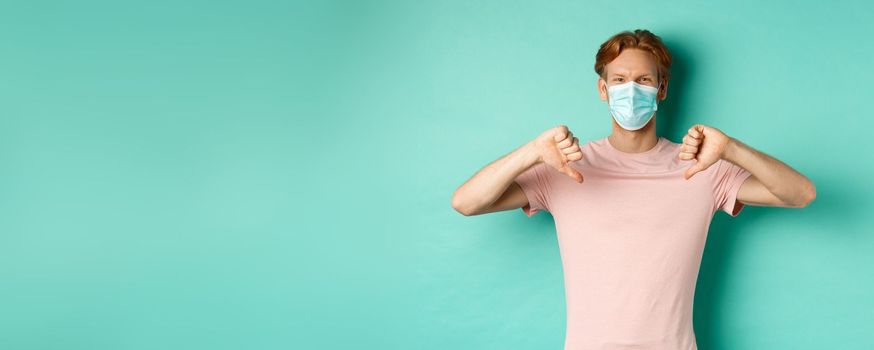 Covid-19, pandemic and lifestyle concept. Young man with red hair in face mask, showing thumbs down, dislike or disapprove something, standing over turquoise background.