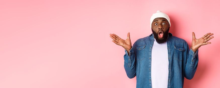 Shocked and impressed Black man staring at camera with complete disbelief, saying wow, standing in beanie and hipster shirt over pink background.