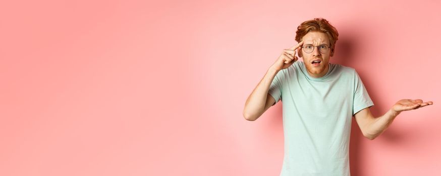 Annoyed redhead man scolding someone stupid, pointing finger at head and shrugging, standing over pink background.