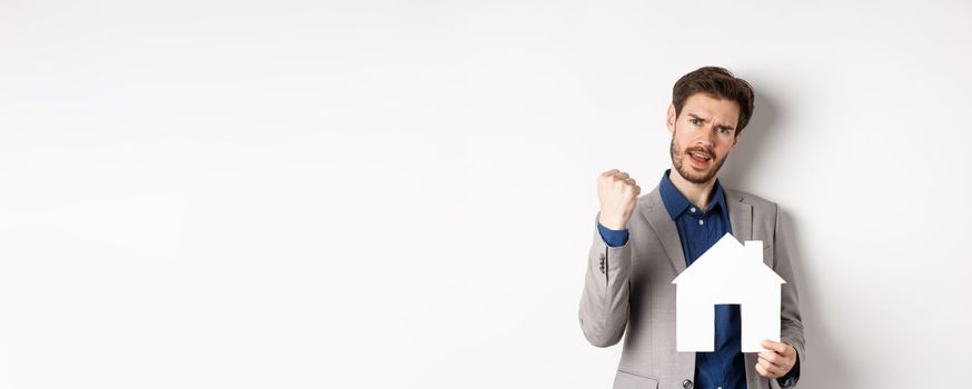 Real estate. Excited man buying house and showing paper home cutout, say yes and fist pump with joy, standing in suit on white background.