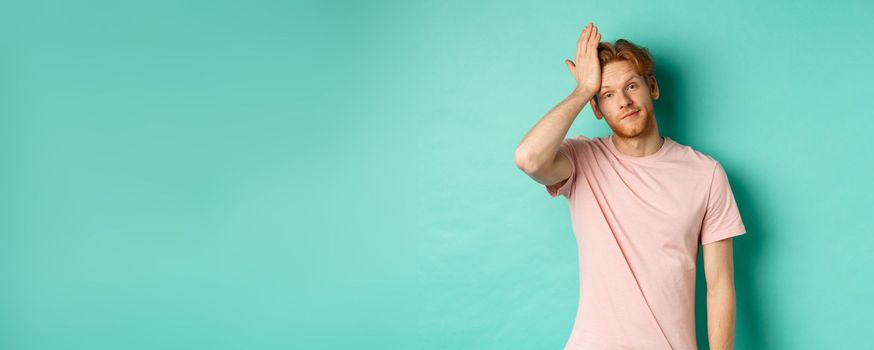 Annoyed and bothered redhead male model showing facepalm gesture, standing over mint background.