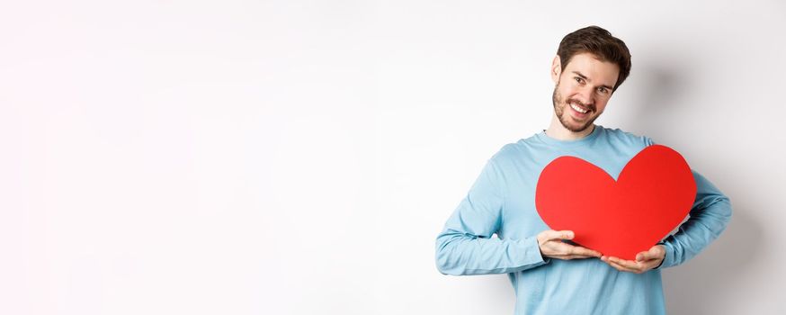 Charming lover man holding Valentines day red heart and smiling, I love you gesture, standing over white background.