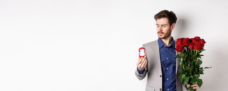 Handsome bearded man in suit looking at engagement ring, making surprise on lovers day, standing with red roses over white background.