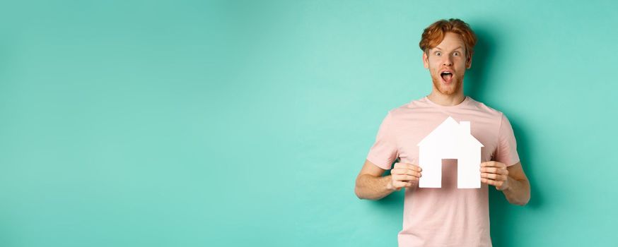 Real estate concept. Excited redhead guy with beard, showing paper house cutout and gasping in awe, staring fascinated at camera, standing over mint background.