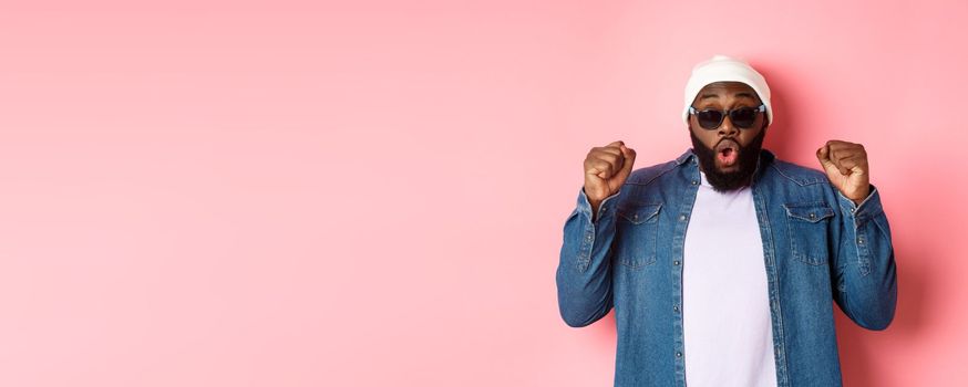 Excited african-american guy in sunglasses and beanie winning and rejoicing, making fist pump in triumph, staring at camera and celebrating, standing over pink background.